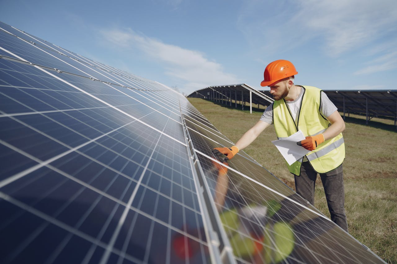 Young bearded male engineer in protective helmet and heat resistant gloves touching solar panel while standing on field with papers on sunny day